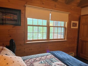 Una habitación tipo cabaña de madera con un ventanal grande, una cama con colcha y un ventilador.