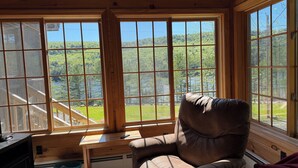 L'intérieur d'une cabane avec une grande fenêtre, un bureau en bois, un fauteuil en cuir et une vue sur une forêt et un lac.