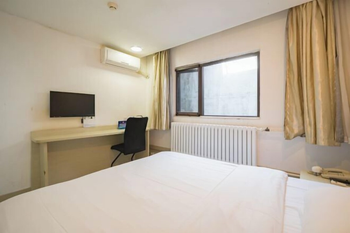 A hotel room with a bed, a desk with a computer, a chair, a window with curtains, and a radiator.