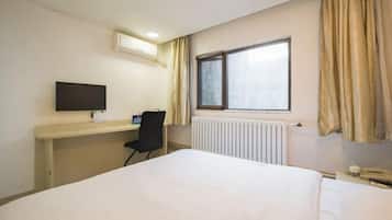 A hotel room with a bed, a desk with a computer, a chair, a window with curtains, and a radiator.