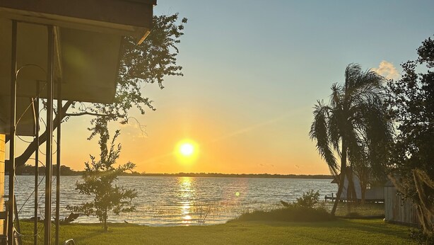 A grassy area with a view of the sunset over a body of water, a covered structure, and a palm tree. - Cozy 1 Bedroom home on Beautiful Lake Alfred (Lake Alfred)