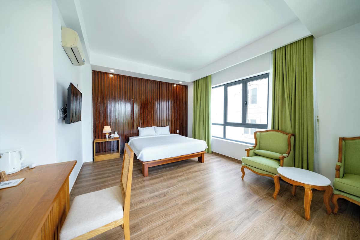 Ein Hotelzimmer mit einem hölzernen Bett, einem grünen Sessel, einem kleinen Tisch, einem Schreibtisch und einem Fernseher.