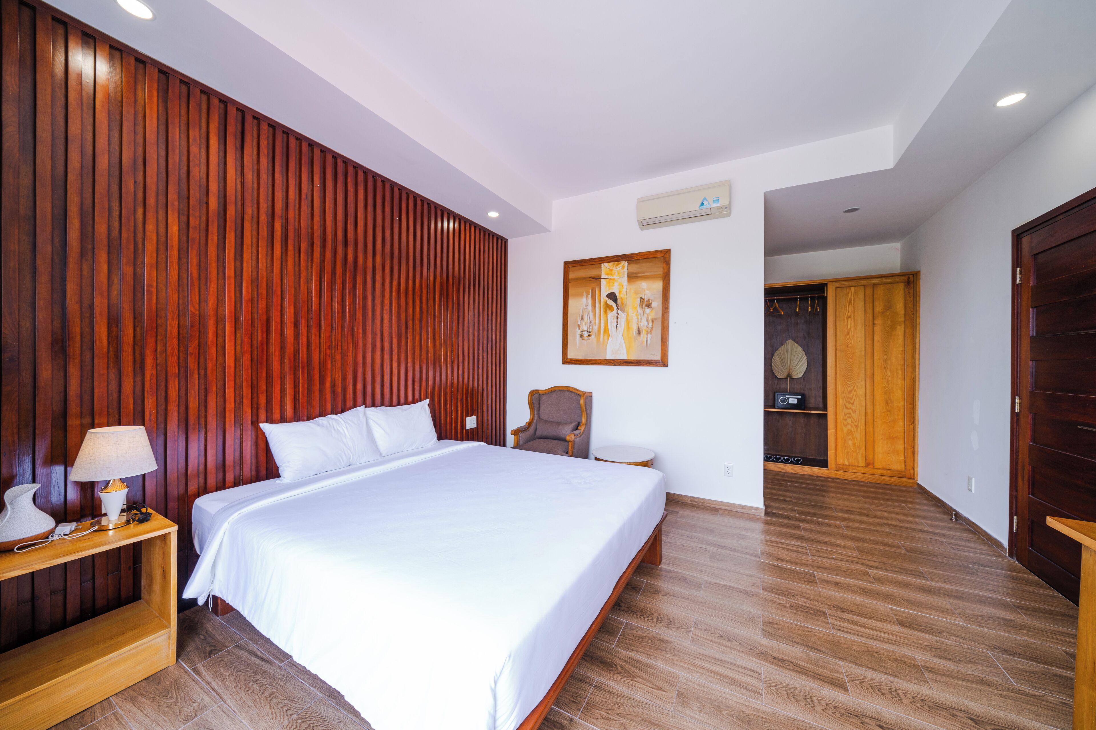 A modern hotel room with a large bed, wooden paneling on one wall, a small table with a lamp, and a painting on the wall.