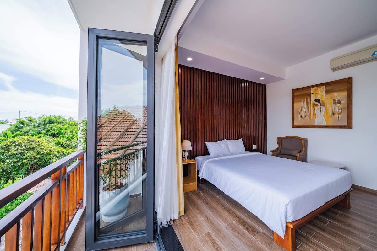 Ein Schlafzimmer mit einem großen Bett, Holzboden, einem Nachttisch, einem Sessel, einem Bild an der Wand und einem Balkon mit Geländer.