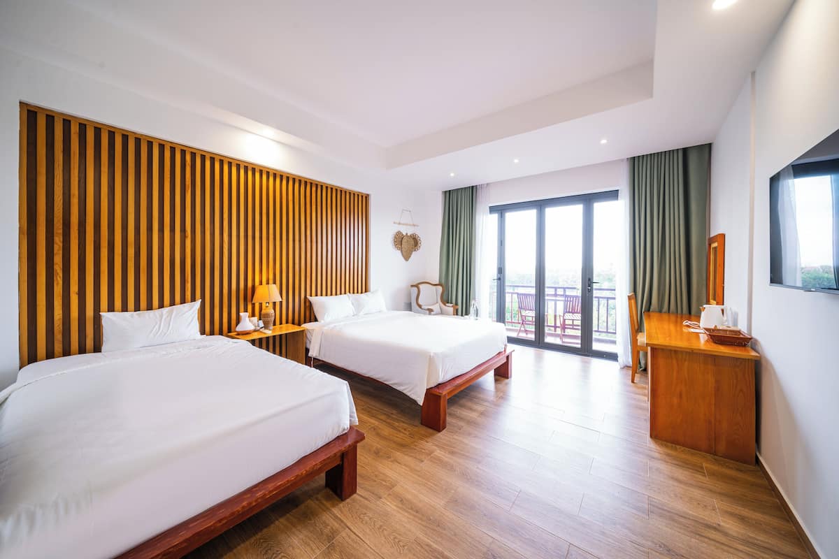 Ein Hotelzimmer mit zwei Betten, einem Holzkopfteil, einem Schreibtisch mit Stuhl und einem Balkon mit Vorhängen.