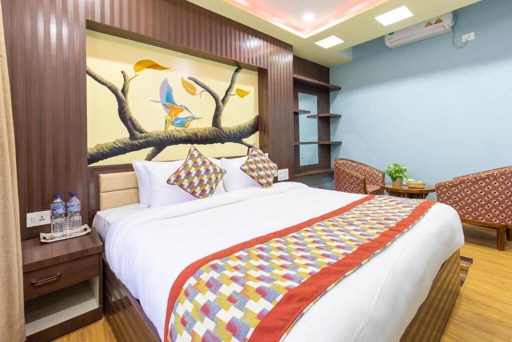 Habitación de hotel con una cama grande, una mesita de noche con botellas de agua, un cuadro de pájaros en un árbol y una silla.