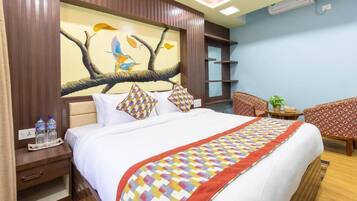 Habitación de hotel con una cama grande, una mesita de noche con botellas de agua, un cuadro de pájaros en un árbol y una silla.