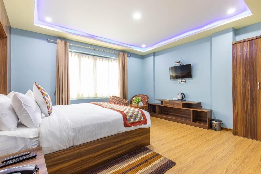 A modern hotel room with a wooden bed, a flat-screen TV mounted on the wall, a small wooden side table, and a window with curtains.