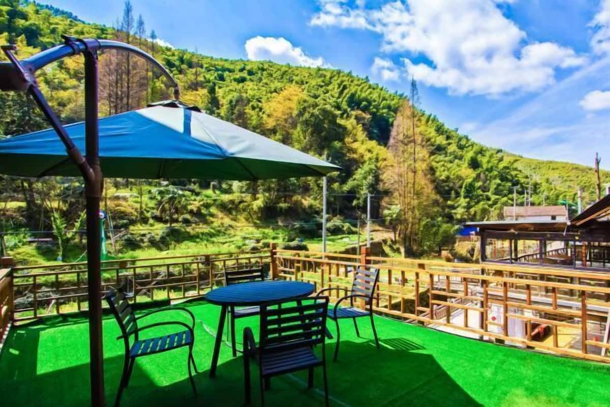 An outdoor seating area with a table and chairs, a wooden fence, and a view of a forested hillside.
