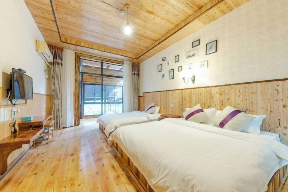 A room with wooden flooring, two beds with white bedding, a desk with a chair, a TV mounted on the wall, and a large window with curtains.