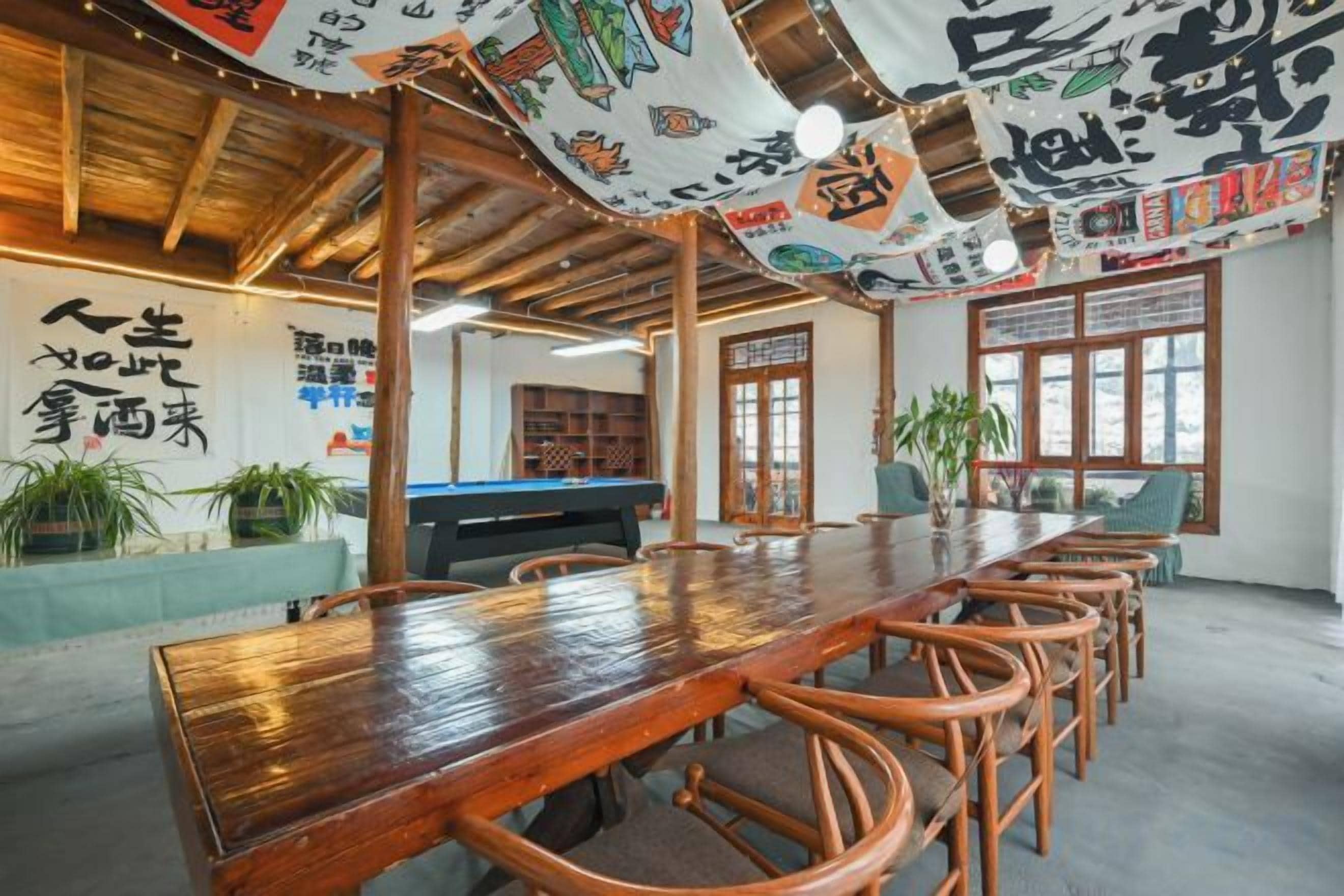 A wooden dining table with chairs, a pool table, and potted plants in a room with a high ceiling and traditional decor.