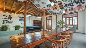 A wooden dining table with chairs, a pool table, and potted plants in a room with a high ceiling and traditional decor.