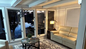 A living room with a sofa, a coffee table with magazines, a lamp, and a large window with a view of trees. - Fountain Blue - Lakeside  (Albert Lea)