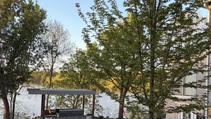 A gazebo with a bench and a table, surrounded by a grassy area and trees, near a body of water and a building. - Fountain Blue - Lakeside  (Albert Lea)