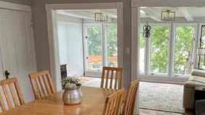A dining area with a wooden table and chairs, a chandelier, and large windows offering a view of greenery. - Fountain Blue - Lakeside  (Albert Lea)