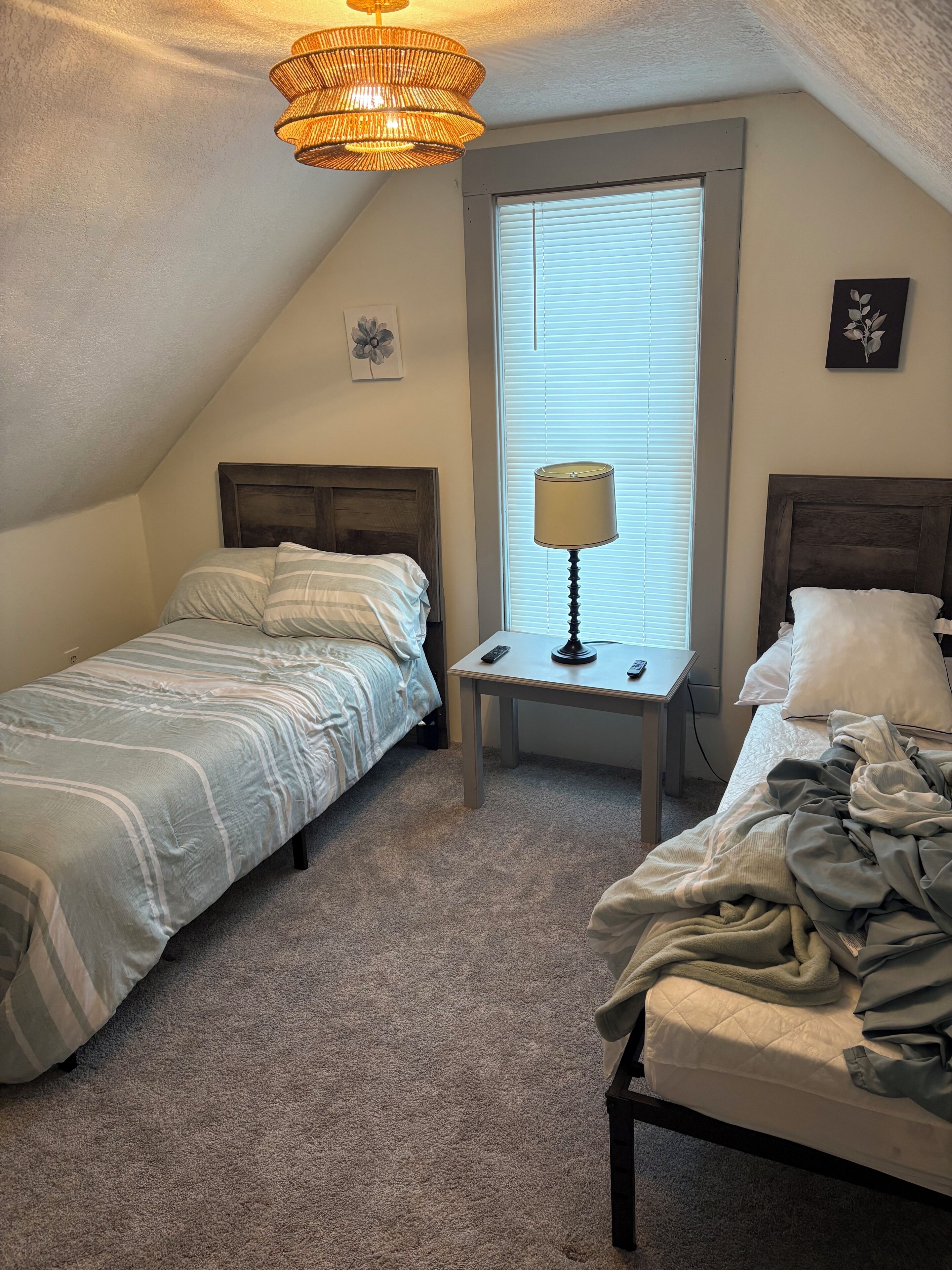 Un dormitorio con dos camas, una ventana con persianas, una lámpara en una mesita de noche y un candelabro.