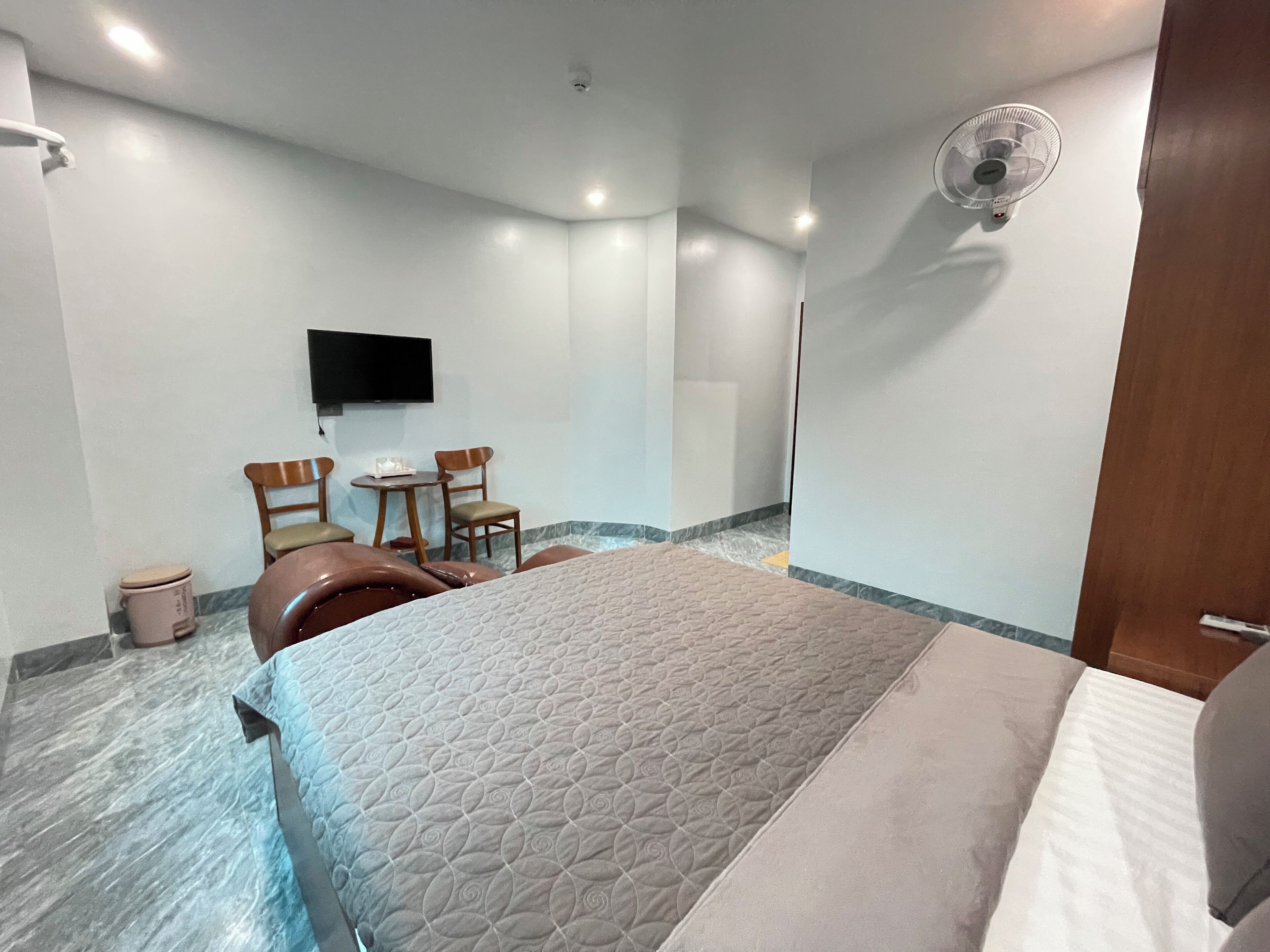 A hotel room with a bed, two chairs, a small table, a TV mounted on the wall, and a ceiling fan.