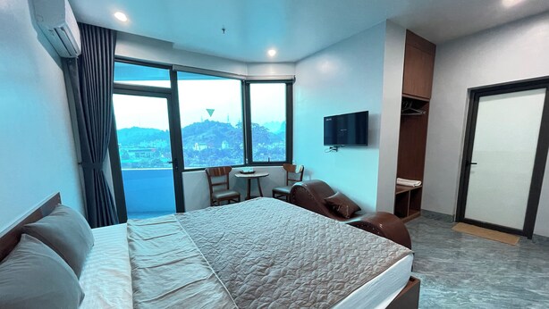 Ein Hotelzimmer mit einem großen Bett, einem kleinen Tisch, einem Sessel, einem Fernseher und Blick auf eine Stadt durch große Fenster.