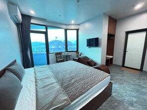 Quarto de hotel com uma cama grande, uma mesa pequena, uma cadeira, uma televisão e vista para a cidade através de grandes janelas.