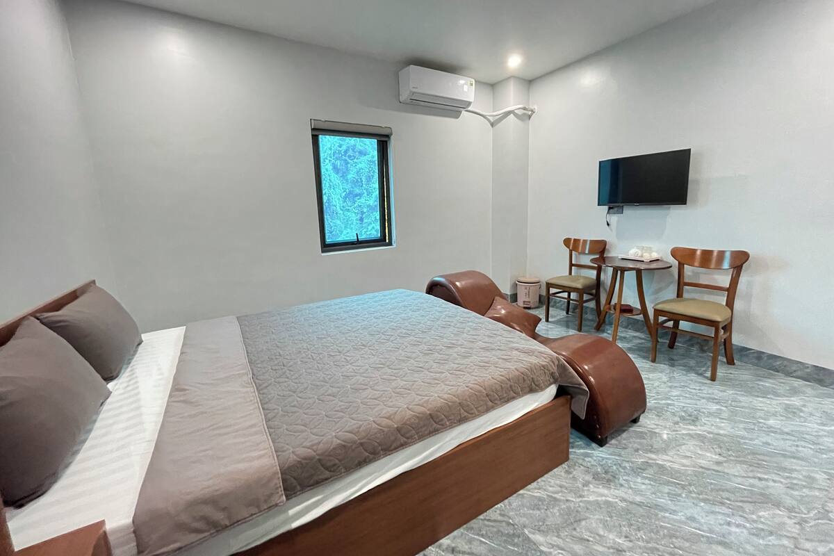 A hotel room with a bed, two chairs, a small table, a TV mounted on the wall, and a window with a view.