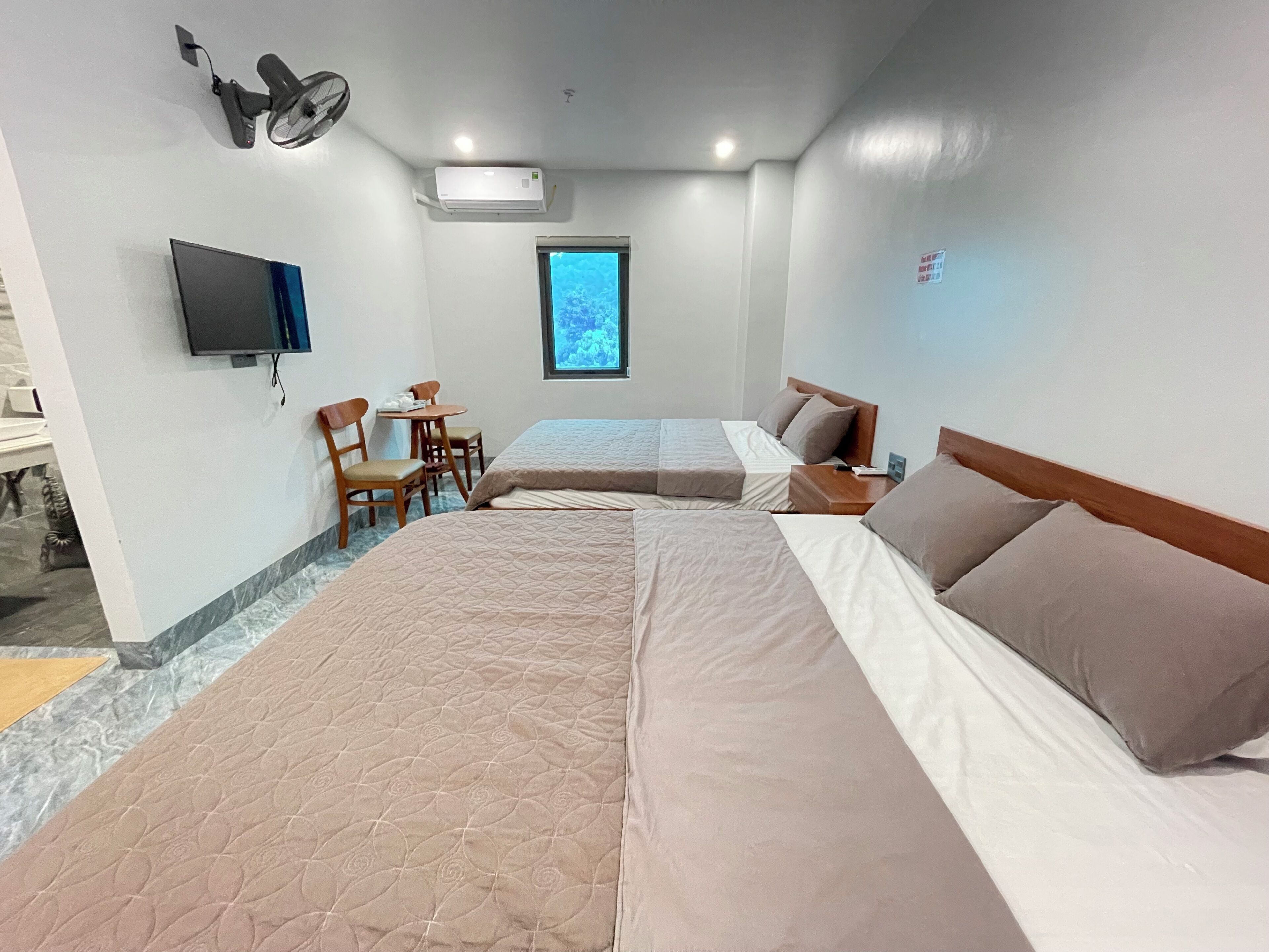 A hotel room with two beds, a desk, a chair, a TV, and an air conditioning unit.