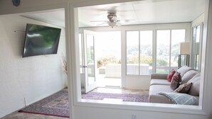 A living room with a flat-screen TV mounted on the wall, a ceiling fan, a sofa with cushions, and a view through large sliding glass doors to an outdoor area.