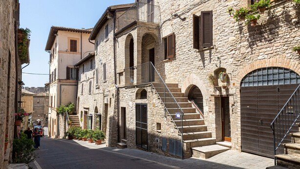 A narrow street with stone buildings, a staircase leading to an upper floor, and a closed metal gate.