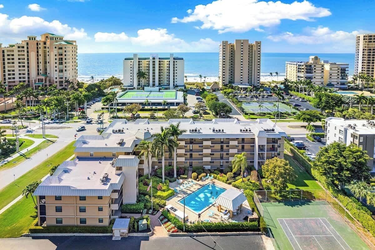 A hotel complex with multiple buildings, a swimming pool, tennis courts, and a view of the ocean.