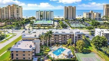 A hotel complex with multiple buildings, a swimming pool, tennis courts, and a view of the ocean.
