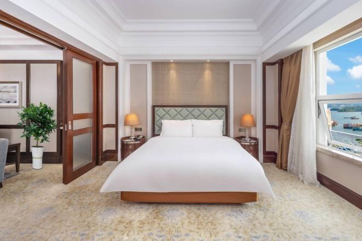 A hotel room with a large bed, two bedside tables, a potted plant, a window with a view, and a door leading to another room.