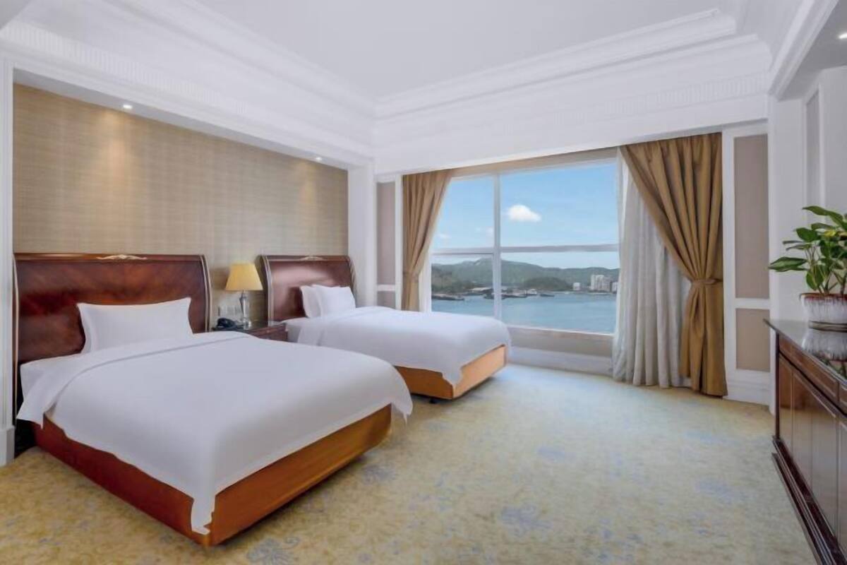 A hotel room with two beds, a large window with a view, and a plant on a side table.