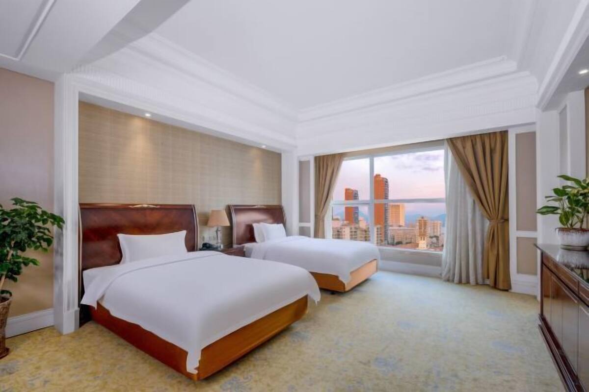 A hotel room with two beds, a large window with a city view, a plant, and a cabinet.