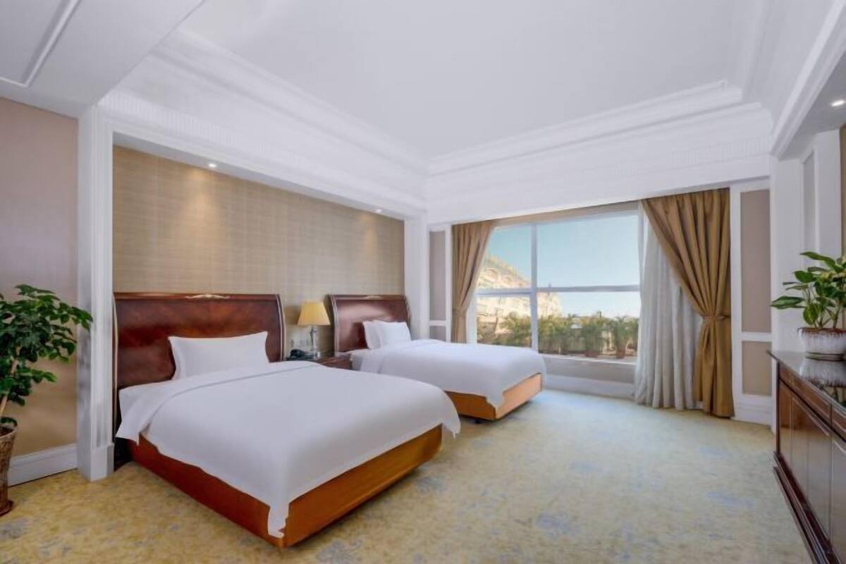 A hotel room with two beds, a large window with curtains, a plant, and a view of the outside.
