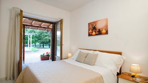 Ein Schlafzimmer mit Bett, Nachttisch, Lampe, einem Gemälde und Blick ins Freie.