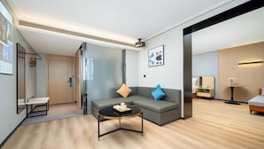 Una habitación de hotel moderna con un sofá gris, una mesita redonda, un escritorio pequeño y una cama con una mesita de noche.