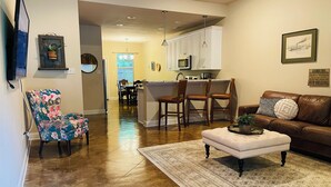 A spacious living room with a brown leather couch, a floral armchair, and a patterned rug. A kitchen area with white cabinets and a bar is visible in the background. - Beech St. fenced back yard, central location, garage (Covington)