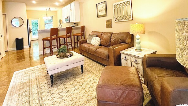 A living room with a brown leather sofa, a matching ottoman, a coffee table, a patterned rug, and a kitchen in the background. - Beech St. fenced back yard, central location, garage (Covington)