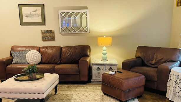 A living room with a brown leather sofa, a matching ottoman, a white tufted coffee table, a patterned rug, a small side table with a lamp, and a framed picture on the wall.
