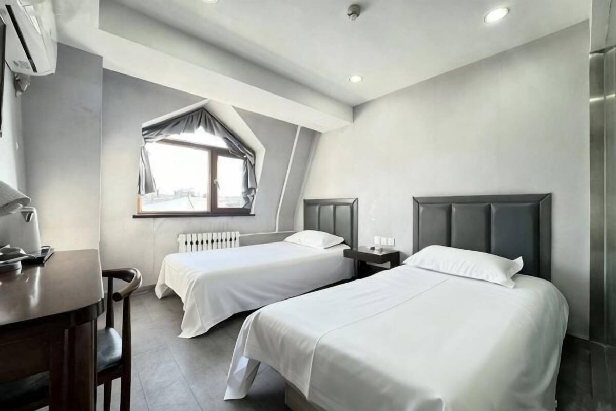 A hotel room with two beds, a desk, a chair, a window with a view, and a radiator.
