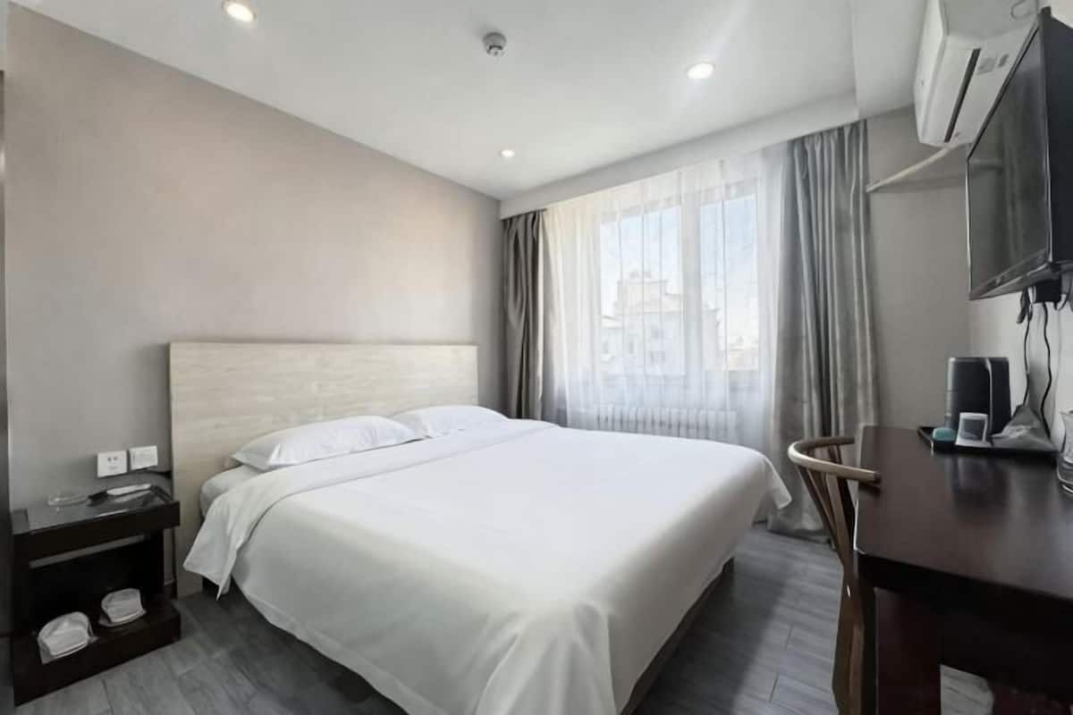 A hotel room with a large bed, a desk with a chair, a flat-screen TV, and a window with curtains.