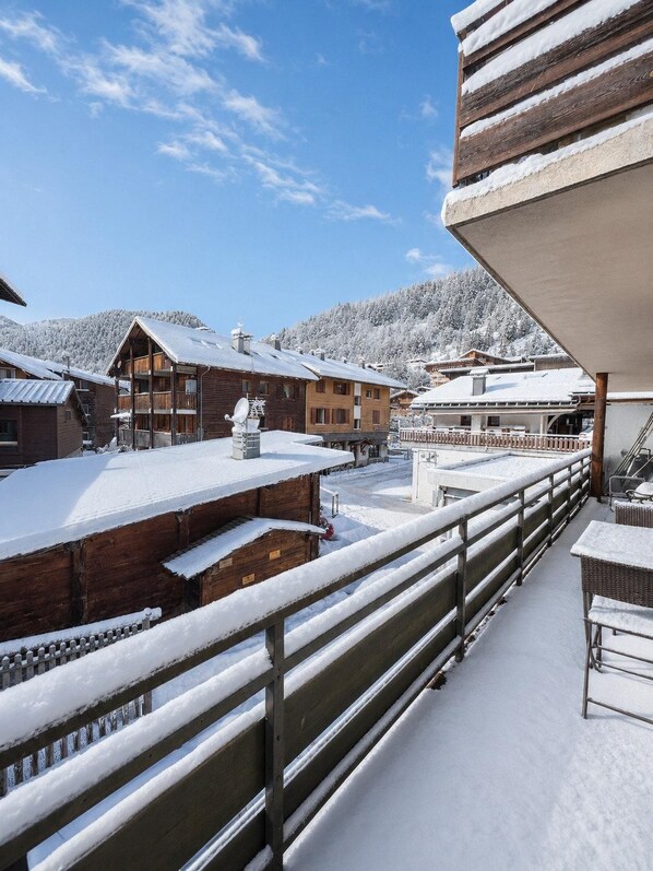 A snow-covered wooden building with a balcony, surrounded by other buildings and a snowy mountain in the background.