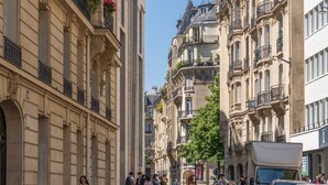 A street lined with historic buildings, featuring balconies with potted plants, a clear sky, and parked cars.