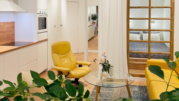 A modern living room with a yellow armchair, a glass coffee table, and a view through a large window with a wooden frame.