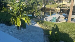 A pathway leading to a dock area with a small boat, surrounded by palm trees and a clear blue sky. - Relaxing 3 Br-2 Ba cottage on canal with heated pool & hot tub. (Cape Coral)