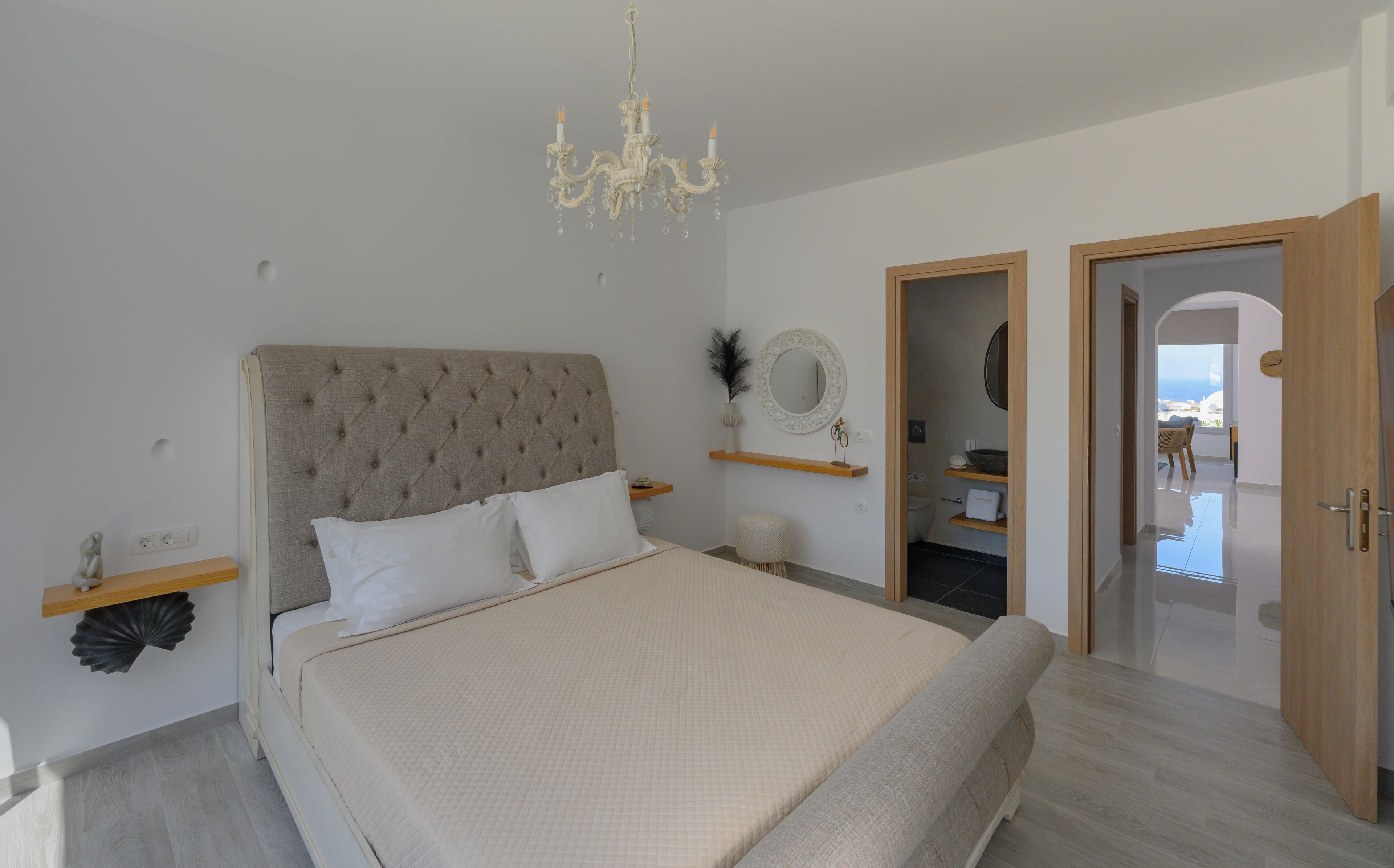 A bedroom with a tufted headboard, a large bed with white bedding, a chandelier, and a view through an open door.