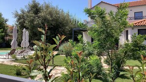 Ein Poolbereich mit Liegestühlen und Sonnenschirmen, umgeben von Grünflächen und einem Gebäude mit einem Ziegeldach.