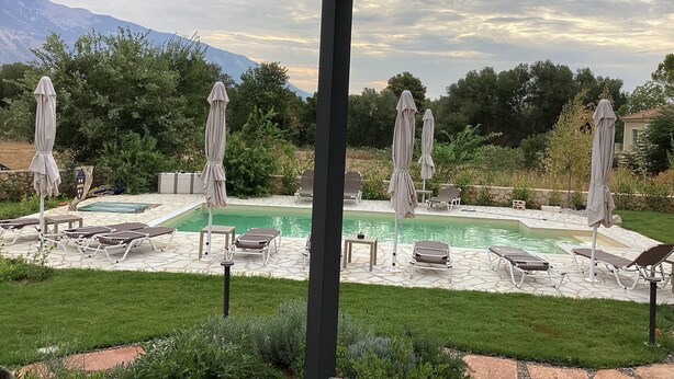 Ein Poolbereich mit Liegestühlen und Sonnenschirmen, umgeben von Grün und Bergen im Hintergrund.