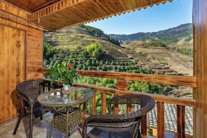 A wooden balcony with wicker furniture, a glass table, and a view of terraced fields.