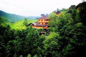 A large, multi-story building with a balcony, surrounded by lush greenery and hills.