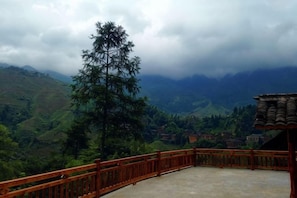 A balcony with a wooden railing overlooking a mountainous landscape with a single tree and distant buildings.